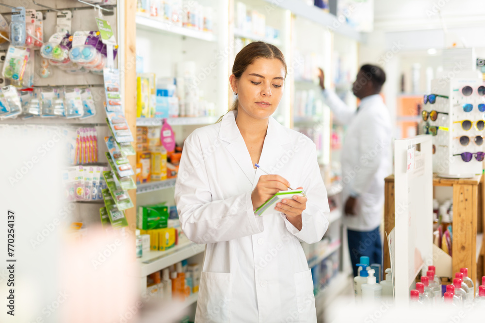 Female pharmacist standing in salesroom of drugstore and holding notebook in hands