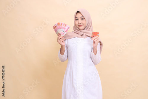 Asian Muslim woman in hijab smiles holding rupiah cash and credit card in hand. Lifestyle, religious, business and finance concepts