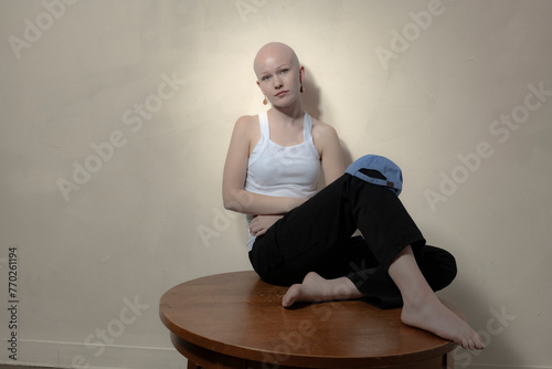 Cuadro en lienzo A bold bald woman with a neutral expression casually holding a blue cap sits on