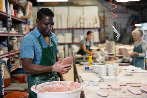 African american worker in pottery factory dips clay plates and cups into a container with glaze