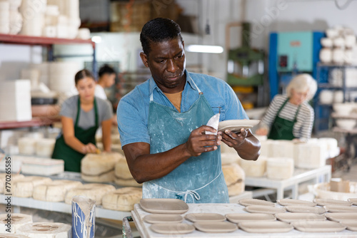 Portrait of african american worker cutting burrs off freshly made earthenware cups and plates with a knife photo