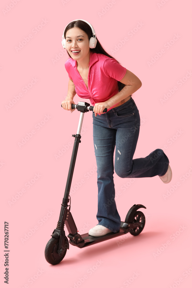 Happy young Asian woman in headphones riding modern electric kick scooter on pink background