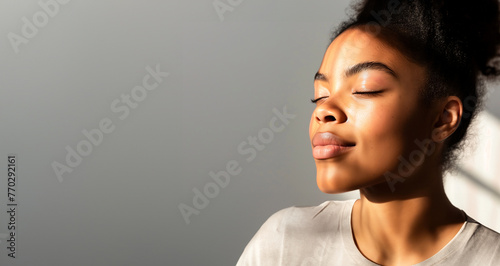 A beautiful african woman with closed eyes is enjoying the sunlight on her face, copy space for text banner on grey background photo