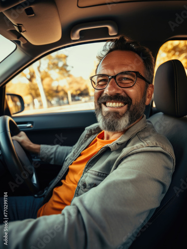 A man in glasses is smiling and driving a car. He is wearing an orange shirt. The car is parked on the side of the road