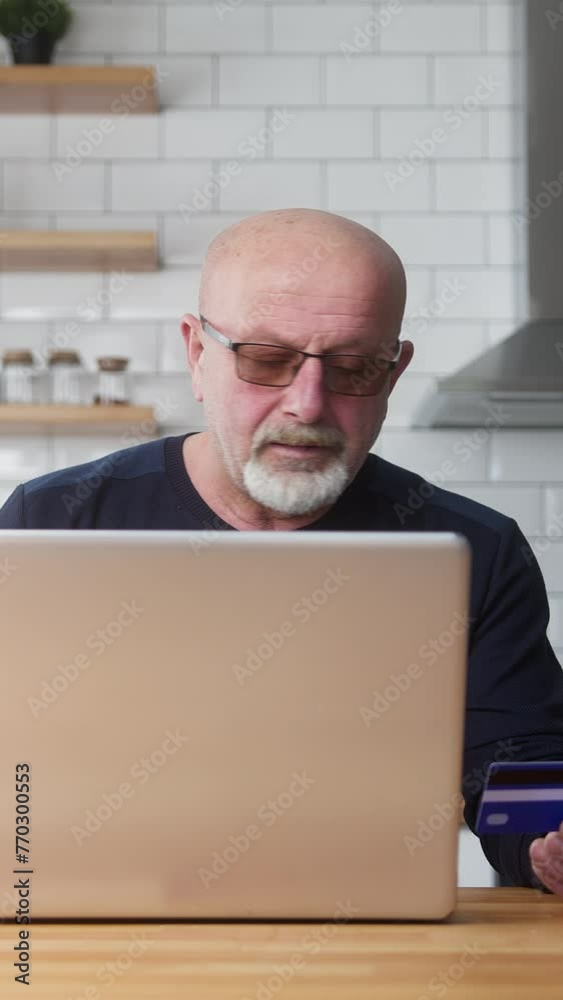 Vertical video. Elderly man enter credit card number on laptop for shopping online, makes secure easy distant electronic payment while sitting in kitchen at home Vertical video. Elderly man enter credit card number on laptop for shopping online, makes secure easy distant electronic payment while sitting in kitchen at home