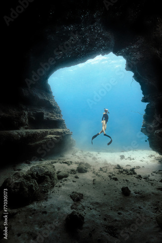 A female free diver leaves an underwater cave opening in the clear blue waters of Hawaii. Model release provided.