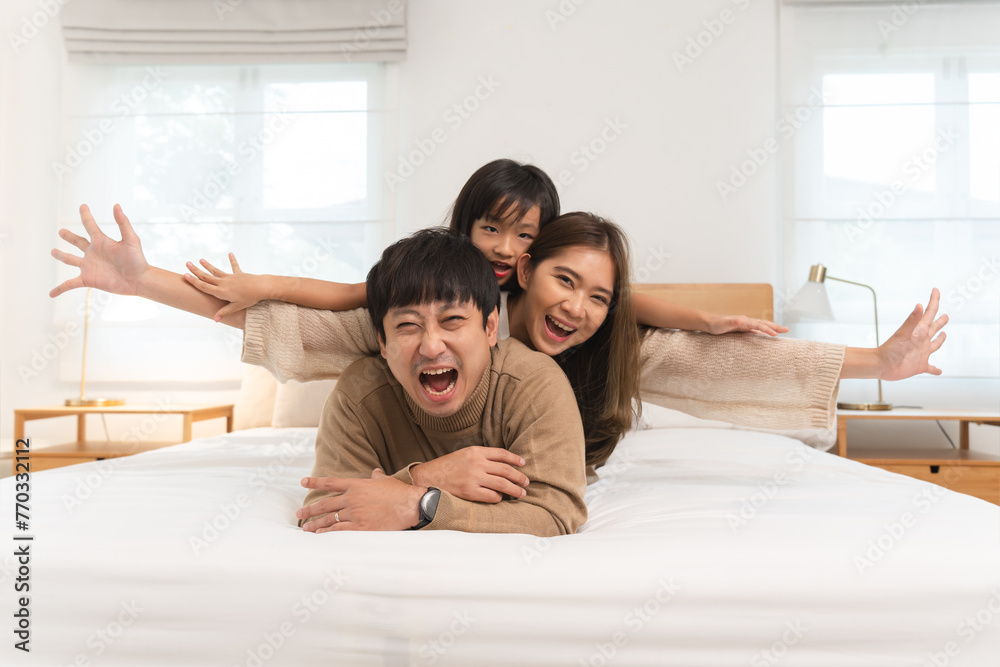 Happy Asian family, young father, mother, cute daughter lying together on bed in bedroom at home. Little girl having fun playing be a plane on her parents smiling with happiness, looking at camera Happy Asian family, young father, mother, cute daughter lying together on bed in bedroom at home. Little girl having fun playing be a plane on her parents smiling with happiness, looking at camera