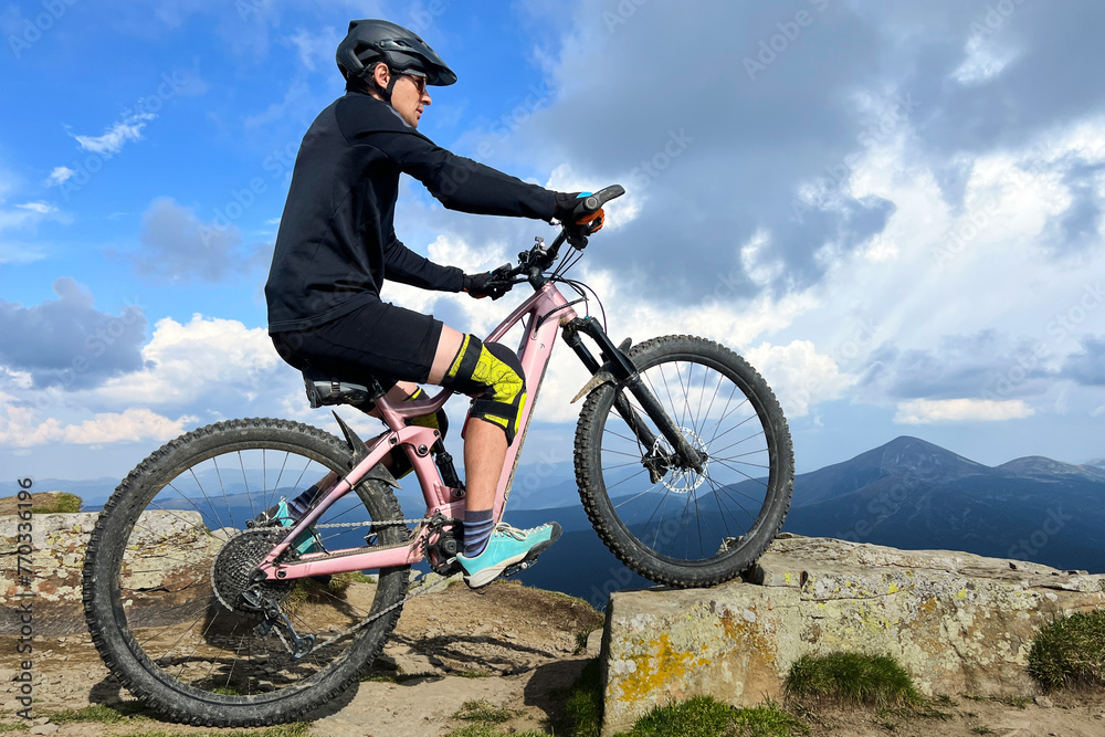 Cyclist man riding electric bike outdoors on sunny day. Male tourist resting on grassy hill, enjoying beautiful mountain landscape, wearing helmet. Concept of active leisure.