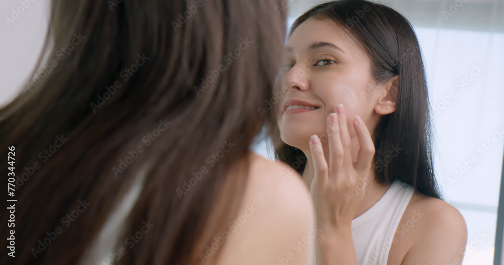 Beautiful young woman applies moisturizer to her face while standing in front of a mirror. Beautiful young woman applies moisturizer to her face while standing in front of a mirror.