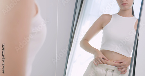 A young woman is worried about her body while looking in the mirror at home. © ronnarong