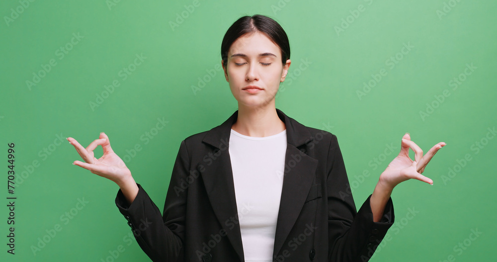 A young businesswoman is relaxing from stress by meditating with her eyes closed. Isolated on green background in studio. A young businesswoman is relaxing from stress by meditating with her eyes closed. Isolated on green background in studio.