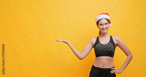 Young sportswoman wearing a Santa hat shows physical strength and points at something. Isolated on yellow background in studio.