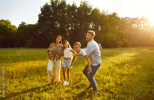 Happy young family of four having fun and playing plane at sunny day in nature. Mother, father and their children boy and girl walking in the park on green grass outdoors at sunset together.