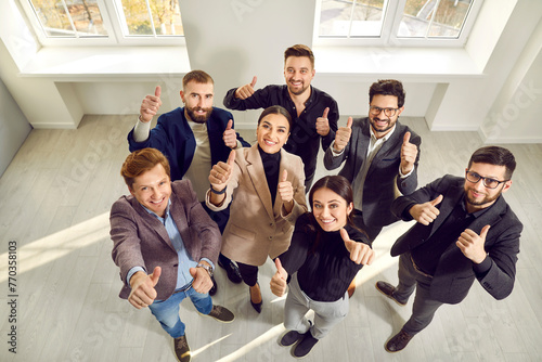 Happy business team giving thumbs up all together. Cheerful young people standing in office, looking up, showing like gestures and smiling. High angle view, shot from above. Success concept