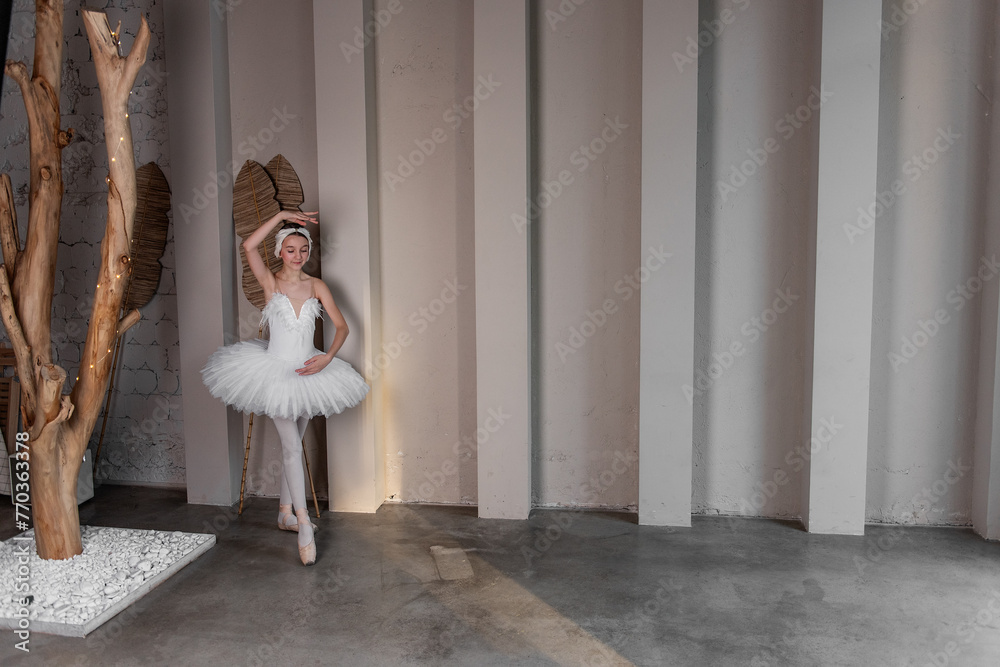 Naklejka premium Elegant young ballerina performs beside large windows, poised stance, delicate white tutu illuminated by natural light, embodying the fusion of strength, grace. White swan performance. Copy space