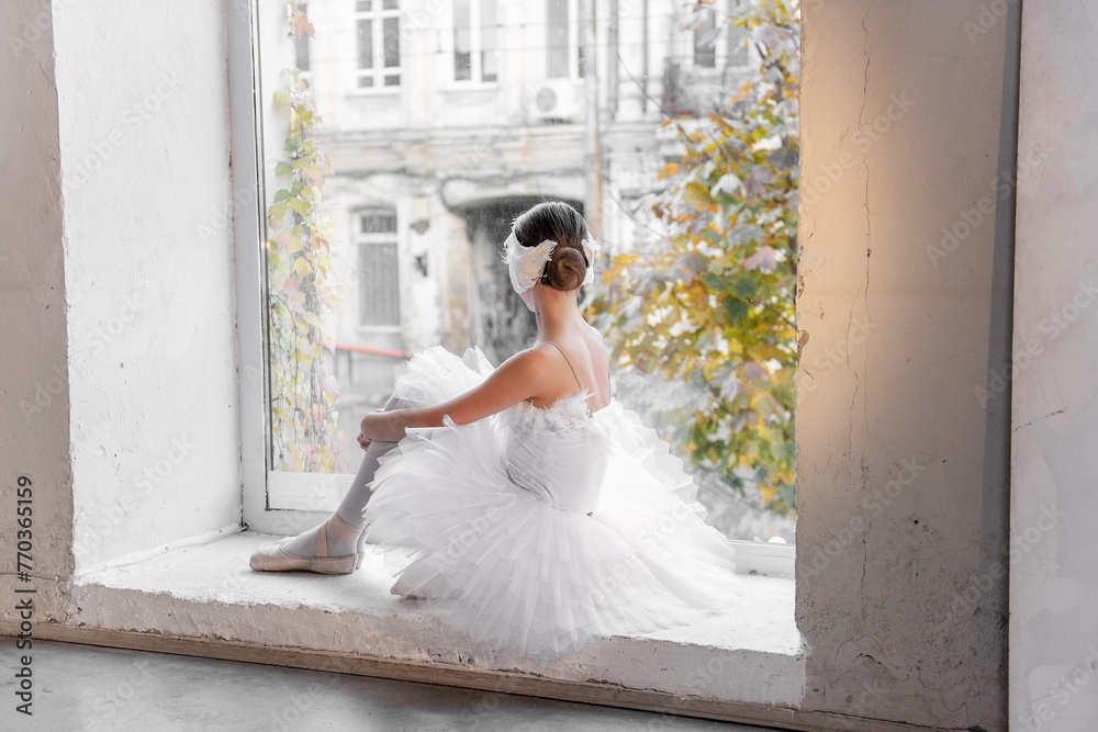 Fototapeta premium Ballerina sits thoughtfully by a sunlit window, her gaze outward, surrounded by urban decay and autumn leaves, in a serene, almost ethereal, moment of stillness.