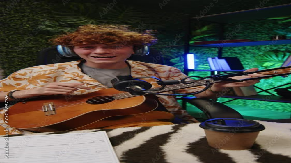 Vertical shot of young cheerful musician in headphones looking at camera, playing guitar and singing, then giving high five to friend while recording content for social media blog in studio Vertical shot of young cheerful musician in headphones looking at camera, playing guitar and singing, then giving high five to friend while recording content for social media blog in studio
