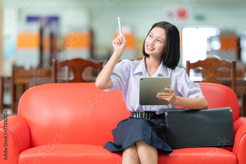 Asian female student with short hair smiles cutely Sitting on the red sofa in the library A pen pointing ahead has a work laptop and a school bag. Wear a uniform in the library to gain knowledge.