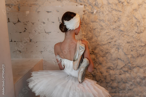 Contemplative ballerina, clad in her feathery white costume, is lost in thought against a textured wall, holding her well-worn pointe shoes in a moment of calm before the performance.