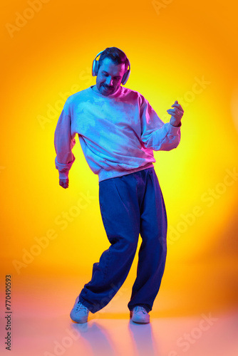 Relaxed, energetic, smiling man in pink sweatshirt and jeans listening to music in headphone and dancing on gradient yellows orange background in neon light. Concept of human emotions, casual fashion