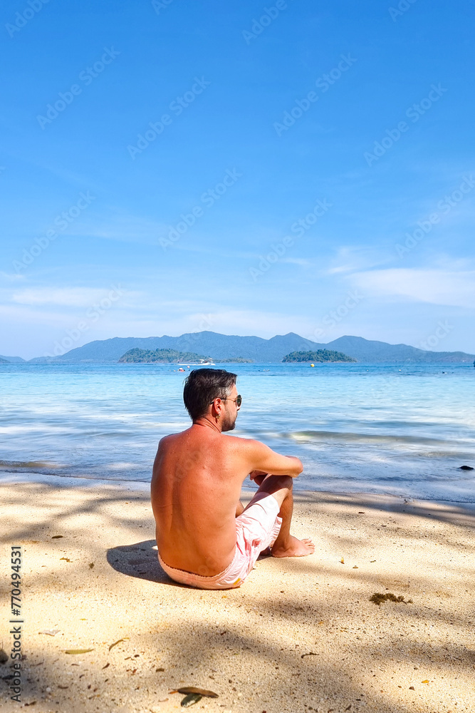 A man in casual clothing sits on the sandy beach, his gaze fixed out to the vast expanse of the ocean before him A man in casual clothing sits on the sandy beach, his gaze fixed out to the vast expanse of the ocean before him