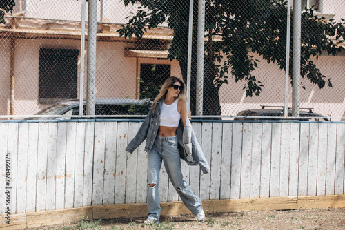 Beautiful blonde model, fashionable in jeans and a denim jacket, poses in the city against the background of a sports field grid photo