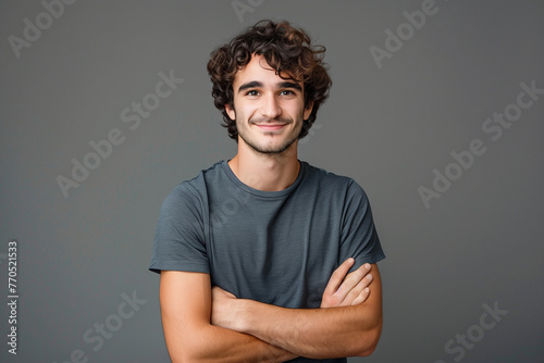 Photo of a happy young man with curly hair in a gray shirt standing on a gray background with his arms crossed and looking at the camera smiling photo