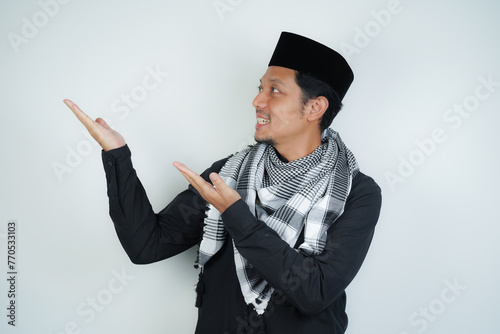 Cheerful handsome Asian Muslim man wearing Arab turban sorban pointing hand finger at empty space for advertising on isolated background photo