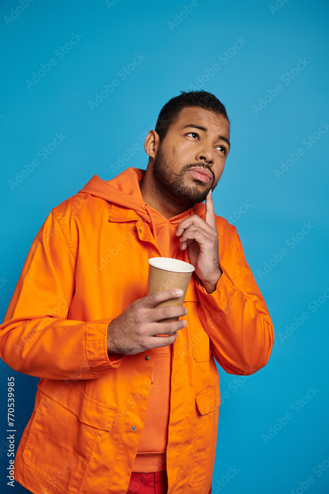 thoughtful african american man in his 20s with paper cup in hand on blue background thoughtful african american man in his 20s with paper cup in hand on blue background