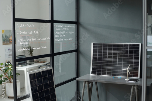 Grey desk with models of solar panel and windmill standing by wall of spacious modern office with workplaces of architects and designers