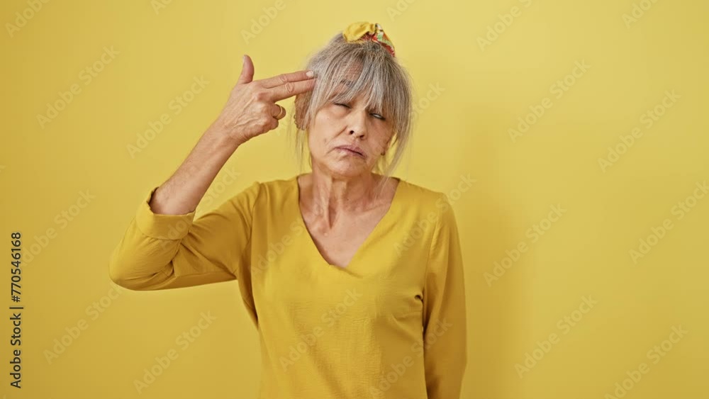 Distraught middle-aged woman with grey hair in a ponytail, shows suicide gesture with her hand mimicking gun, pointing to head in isolated yellow background. Distraught middle-aged woman with grey hair in a ponytail, shows suicide gesture with her hand mimicking gun, pointing to head in isolated yellow background.