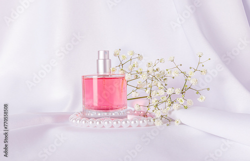 Glass bottle of pink perfume on white satin background with gypsophila branch and pearl beads. The concept of natural floral scents. Front view.