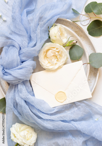 Envelope near light blue fabric knot and cream roses on plates top view copy space, wedding mockup