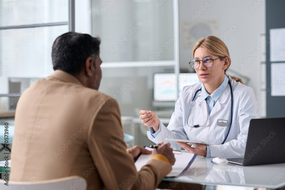 Fototapeta premium Portrait of smiling female doctor consulting Middle Eastern man sitting across table in medical clinic