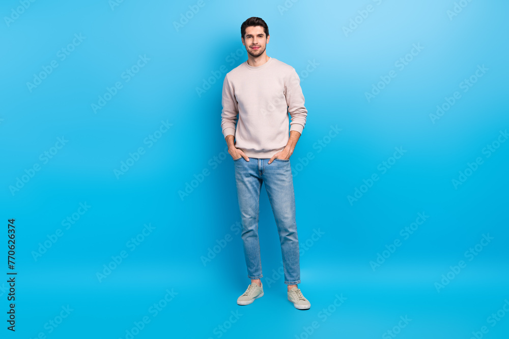 Full size photo of nice young man put hands pockets wear beige sweatshirt isolated on blue color background Full size photo of nice young man put hands pockets wear beige sweatshirt isolated on blue color background