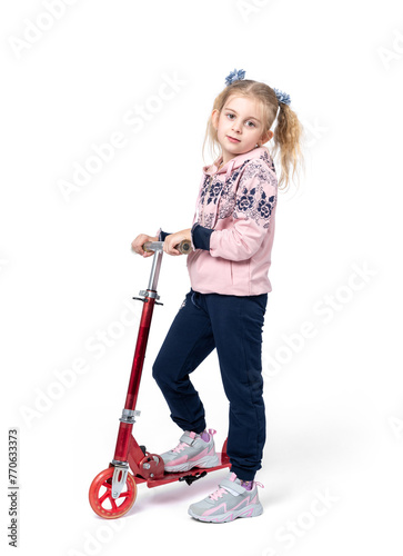 A happy little girl in sports clothes stands with a red scooter, isolated on a white background