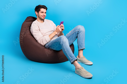 Full size photo of clever positive man wear sweatshirt denim pants sit on bean bag look at smartphone isolated on blue color background © deagreez