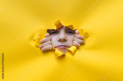 A little phlegmatic girl looks into a torn hole in yellow paper. The concept of espionage, childish curiosity, peeping, indifference to what is seen. Copy space.