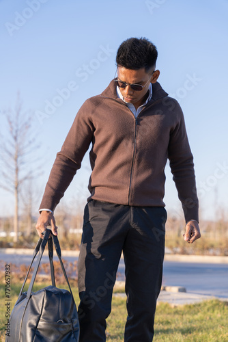 A man wearing a brown jacket and black pants is holding a black bag. He is standing in a grassy area with a blue sky above him. Concept of casualness and leisure © oybekostanov