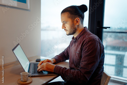 Side portrait of serious handsome man with hair bun  working at laptop, typing on keyboard, smartphone and espresso coffee on table, background of panoramic window.