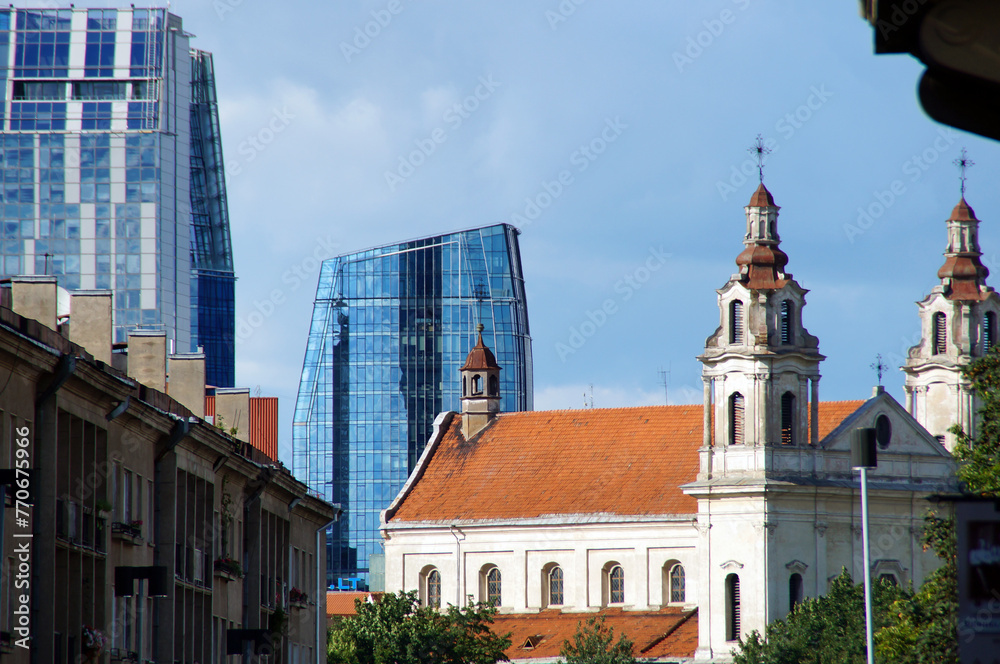 Obraz premium An old religious building, a cathedral with bright orange roof tiles, towers, columns, against which the blue sky and tall buildings belonging to modern architecture with glass facades
