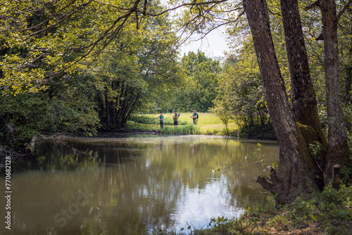 Three sports fishermen standing on the river bank, under a beautiful tree, swinging their fishing rods trying to catch fish on a sunny summer day.