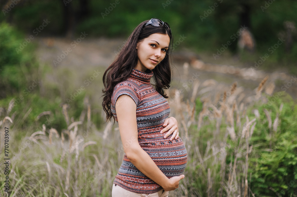 A beautiful smiling young pregnant brunette woman stands in nature, outdoors, holding her big belly with her hands. Pregnancy photography, portrait. A beautiful smiling young pregnant brunette woman stands in nature, outdoors, holding her big belly with her hands. Pregnancy photography, portrait.
