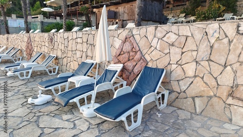 Plastic lounge sunbed with soft mattresses, umbrellas and tables are arranged on a tiled area near the wall. Palm trees grow nearby Everything is illuminated by the soft sunset light of the autumn sun
