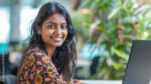 Close-up portrait of a smiling young Sri Lankan woman with long black hair seated and working on a laptop