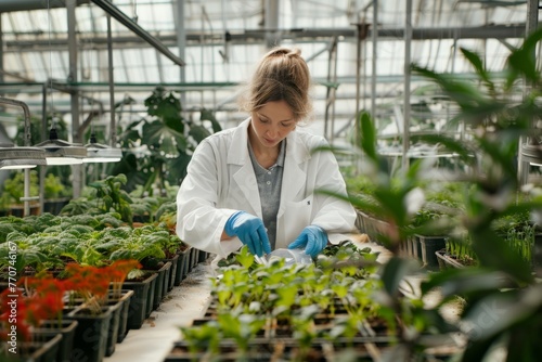 A woman wearing a white coat and gloves is actively working inside a greenhouse at a botanical research facility