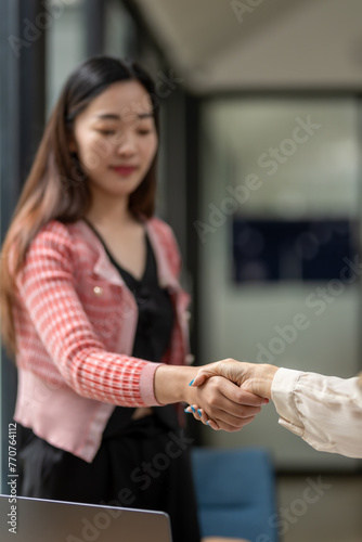 Two professional women in a modern office setting confidently shake hands over a successful business agreement or partnership.