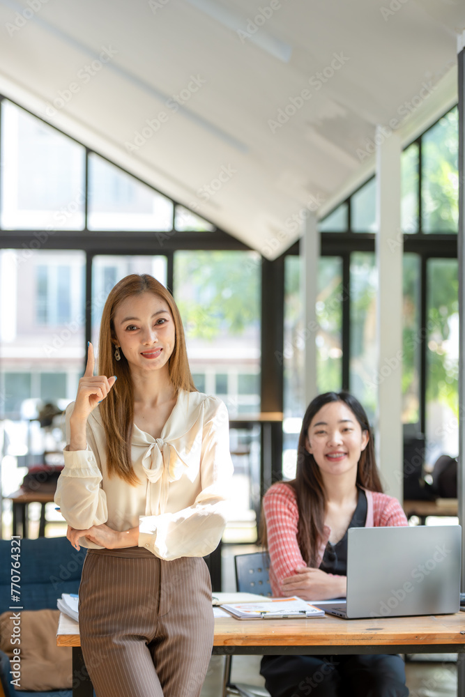 A confident businesswoman stands arms crossed in a modern collaborative workspace with a seated colleague in the background. A confident businesswoman stands arms crossed in a modern collaborative workspace with a seated colleague in the background.