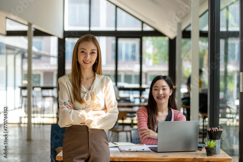 A confident businesswoman stands arms crossed in a modern collaborative workspace with a seated colleague in the background.