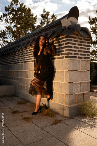 A dramatic shot of a woman in a black fringed dress and heels, leaning against a traditional Korean-style wall under the soft glow of sunset.